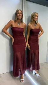 Vestido largo color bordó, de estilo strapless con escote recto. Presenta un cuerpo ajustado con detalles de fruncido en la zona de la cintura y cadera, y una falda inferior confeccionada en tela semitransparente con diseño plisado.