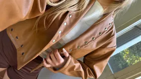 Campera corta de ecocuero color camel, con diseño de cuello solapa y cierre frontal con botones metálicos a presión. Presenta detalles de tachas metálicas a lo largo de la solapa y en el ruedo.