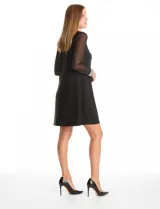 Vestido negro de seda fría con silueta evasé, cuello redondo y mangas largas de gasa transparente. Los puños son anchos y están decorados con una densa aplicación de perlas blancas.