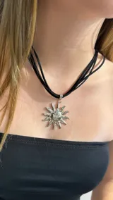 Gargantilla tipo choker compuesta por múltiples tiras finas de gamuza negra, con un dije central metálico en forma de sol con rostro grabado.