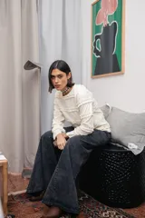 Blusa de color manteca con textura acanalada, cuello redondo y mangas largas con puños acampanados. El diseño presenta múltiples volados horizontales distribuidos a lo largo del cuerpo y las mangas.