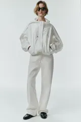 Chaqueta blanca de cuello redondo con capucha ajustable mediante cordones elásticos. Presenta manga larga con puños elásticos, bolsillos delanteros y bajo asimétrico ajustable con cordones y stoppers. El cierre frontal combina cremallera y botones ocultos bajo una solapa.
