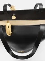 Bolso tipo tote asimétrico de cuero liso color negro, con diseño de cubo, doble asa superior y cierre de anilla metálica dorada. Incluye logo de la marca en letras doradas en la parte inferior.