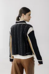 Chaqueta de punto con diseño color block en negro, beige y marrón. Presenta cuello redondo, cierre frontal con botones grandes, bolsillos frontales y detalles de costuras decorativas verticales en color claro.