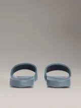 Ojotas tipo slide de Calvin Klein Jeans en color azul, con logo de la marca en relieve en color verde neón sobre la banda superior y logo grabado en el lateral de la suela.