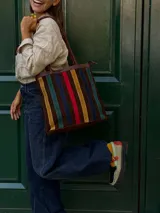Cartera tipo tote de cuero con diseño de franjas verticales multicolores en tonos tierra, rojo y azul. Cuenta con correas trenzadas de cuero y cierre superior.