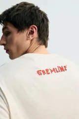 Camiseta de corte relajado color crudo, con cuello redondo y manga corta. Presenta un estampado frontal con una ilustración de Gizmo de la película Gremlins conduciendo un auto rojo y el texto 'Never ever feed them after midnight'.