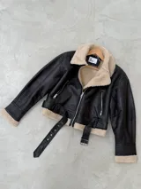 Campera tipo gamulán de cuero sintético negro con interior y cuello de corderito beige. Presenta cierre frontal, bolsillos laterales con cierre y cinturón ajustable en la cintura con hebilla metálica.