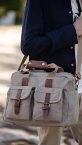 Bolso tipo maletín de tela canvas color beige con detalles en cuero sintético marrón. Cuenta con dos bolsillos frontales con tapa y cierre de hebilla, cierre superior, doble asa de mano y correa larga ajustable y desmontable.