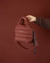 Bolsito tipo tote acolchado de color amarillo, con diseño de líneas horizontales y logo de la marca estampado en el frente. Cuenta con dos asas superiores acolchadas y una correa ajustable de cinta en color beige.