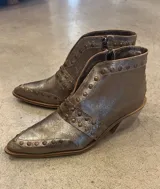 Botín corto de cuero color plata metalizado, con punta fina y cierre lateral. Presenta tachas metálicas decorativas en el empeine y en una tira que rodea el tobillo. Tiene un taco bajo y suela de color natural.