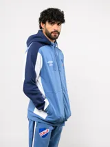 Campera deportiva Umbro del Club Nacional de Football, con cierre frontal, capucha ajustable con cordones y diseño en bloques de color azul, celeste y blanco. Presenta el logo de la marca y el escudo del club bordados en el pecho.