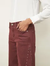 Jean de corte recto y tiro medio en color bordeaux, con detalle de tachas metálicas a lo largo de las costuras laterales y ruedo desflecado.
