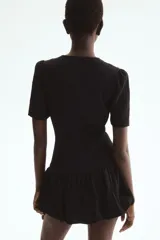 Vestido corto negro con escote redondo, mangas cortas abullonadas y cierre frontal de botones forrados. Cuenta con costuras moldeadoras en la cintura y falda con bajo globo fruncido.