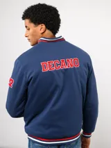Campera bomber azul marino estilo retro, con cierre frontal de botones a presión, cuello, puños y ruedo con tejido acanalado tricolor. Presenta el logo de Umbro y el escudo del Club Nacional de Football bordados en el pecho, junto con el año 1899 y parches decorativos en las mangas.