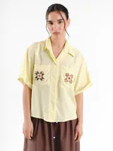 Camisa de manga corta color amarillo claro, con calce oversized, hombros caídos y cierre frontal con botones. Cuenta con bolsillos delanteros con bordados geométricos y mangas con dobladillo.