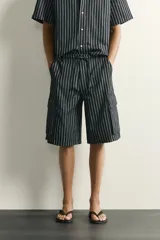 Bermuda cargo relaxed fit confeccionada en denim de mezcla de algodón y lino, con diseño de rayas verticales grises y negras. Cuenta con bolsillos laterales tipo cargo con solapa y cierre frontal con cremallera y botón.