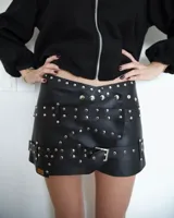 Minifalda negra de cuero sintético con diseño cruzado, decorada con múltiples tachas metálicas plateadas y una correa ajustable con hebilla en la parte inferior.