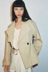 Trench corta de color beige con cuello de solapa y manga larga. Cuenta con bolsillos delanteros, cintura ajustable con cordones, bajo abullonado y cierre frontal cruzado con un botón visible. El interior está forrado con tela a cuadros.