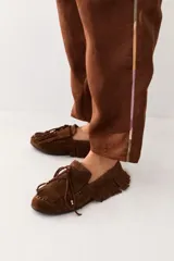 Mocasín de gamuza color marrón, con diseño de flecos decorativos en todo el contorno y la capellada. Presenta cordones decorativos con detalles metálicos dorados en las puntas.