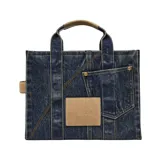 Bolso tipo tote de Marc Jacobs confeccionado en denim, con diseño estructurado que incluye dos bolsillos frontales con solapa y cierre de broche metálico. Presenta el logo bordado en el frente y asas superiores reforzadas junto con una correa ajustable para llevar al hombro.