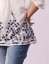 Blusa blanca de gasa bambula con cuello a la base, escote en V y pliegues. Tiene apertura central con botones, mangas ¾ con terminación fruncida y ruedo con detalles bordados en negro.