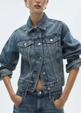 Chaqueta de jean estilo vaquero con cuello camisero, cierre frontal de botones metálicos, dos bolsillos de solapa en el pecho y mangas largas con puños abotonados.