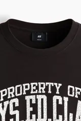Remera negra de algodón con cuello redondo, manga corta y hombros caídos, de corte holgado. Presenta un estampado gráfico frontal estilo universitario en color blanco con efecto desgastado que lee "PROPERTY OF PHYS. ED. CLASS 1905 STATEN ISLAND".
