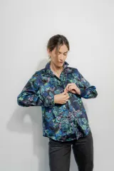 Camisa de seda con cuello clásico y cierre frontal de botones, de manga larga con puños abotonados. Presenta un estampado floral en tonos azules, verdes y rosados sobre fondo negro.