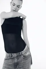 Body sin mangas de cuello redondo, confeccionado en una combinación de tul transparente y paneles de tela opaca a tono. Presenta un diseño ajustado con cierre inferior mediante botones a presión.