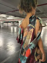 Vestido largo de satén sublimado con diseño abstracto multicolor. Tiene un hombro descubierto y un gran lazo decorativo.