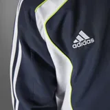 Campera deportiva Adidas del Real Madrid, color azul marino con detalles en blanco y amarillo. Presenta cierre frontal, cuello alto, bolsillos laterales, puños elásticos, dobladillo ajustable y escudo del club bordado en el pecho.