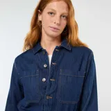 Mono de jean azul oscuro con cierre frontal de botones, cuello camisero, mangas largas y bolsillos frontales. Presenta un corte recto y holgado en las piernas.