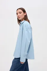 Camisa de algodón en color celeste claro, con cuello solapa, cierre frontal de botones y bordado pequeño en el pecho. Presenta un corte holgado y mangas largas.