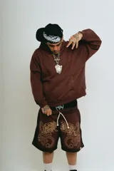 Bermuda de felpa color marrón oscuro con efecto lavado (washed) y detalles en negro en los laterales. Presenta un gran estampado serigrafiado en dorado con tipografía oriental y motivos ornamentales. Tiene cordón de ajuste en la cintura y remaches metálicos cromados en los laterales.