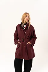 Trench de corte holgado color bordó, con cuello camisero, cierre frontal con botones y cinturón ajustable con hebilla. Presenta mangas largas con detalles de correas en los puños y bolsillos laterales.