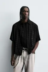 Camisa negra de corte relajado con tejido semitransparente de estructura abierta y patrón a cuadros. Presenta cuello solapa, manga corta, bolsillo plastrón en el pecho y cierre frontal de botones.