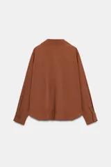 Camisa de lino color beige, con cuello solapa, escote en V y mangas largas. Presenta un diseño de bajo asimétrico con pliegues delanteros que generan un efecto abullonado.