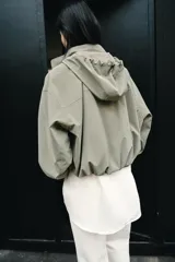 Cazadora de cuello subido con capucha ajustable, manga larga con puños elásticos y bolsillos delanteros con solapa. Presenta bajo elástico y cierre frontal con cremallera oculta y botones a presión.