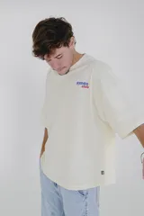 Remera color crema de corte holgado, con cuello redondo y estampado pequeño en el pecho con la frase 'Karma Club' en azul y rojo.