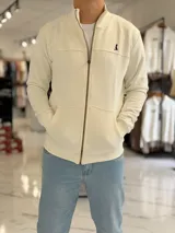 Campera de felpa color beige con cierre frontal, bolsillos tipo canguro y cuello alto estilo bomber. Cuenta con un pequeño bordado en el pecho.