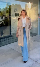 Trench coat color beige de corte clásico, con solapas amplias, doble botonadura frontal, cinturón ajustable con hebilla y trabillas en los puños.