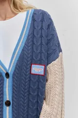 Cardigan de punto con diseño de bloques de color, combinando azul marino en el cuerpo y beige en las mangas. Presenta un tejido con textura de trenzas, cuello en V con borde rayado en azul y blanco, cierre frontal con botones y bolsillos delanteros. Incluye un parche decorativo en el pecho.