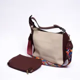 Bolso tipo hobo color beige con detalles en marrón. Tiene correa de mano marrón y correa bandolera ancha de tela estampada multicolor. Incluye un monedero marrón con cierre.