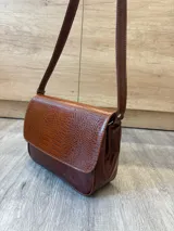 Cartera bandolera de cuero vacuno color bordó, con solapa frontal de textura símil cocodrilo y detalles de costuras pespunteadas en blanco. Cuenta con correa ajustable y cierre superior.