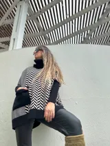 Poncho oversize con cuello alto negro, diseño geométrico en blanco y negro, bolsillo lateral y detalle de bloques de color en gris y negro.