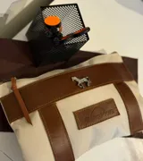 Sobre tipo cartuchera marca Sierra Mora, confeccionado en lona color beige con detalles y terminaciones en cuero marrón. Presenta un aplique metálico en forma de caballo y un parche de cuero con el logo de la marca grabado.