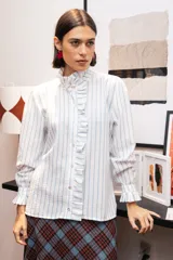 Camisa de manga larga con estampado de rayas verticales celestes sobre fondo blanco. Presenta un cuello alto con volados, cierre frontal con botones y puños con volados.