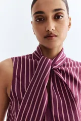 Blusa sin mangas color morado oscuro con finas rayas verticales en color claro. Presenta un cuello alto que se anuda al frente formando un lazo grande.