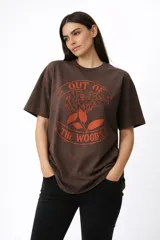 Remera de algodón color marrón con estampado frontal en color naranja que incluye motivos florales y la frase 'Out of the woods'. Presenta cuello redondo y diseño de corte holgado.